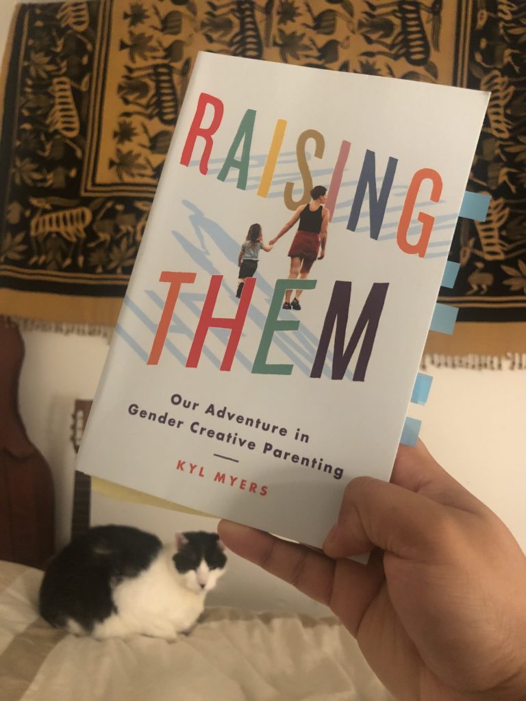  hand holding the book "Raising Them by Kyl Myers" with Kitty B underneath and Guitar handle behind them 